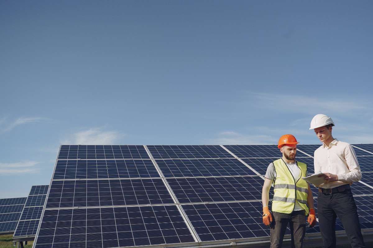 Twee mannen in veiligheidshelmen staan voor een rij zonnepanelen, met een heldere lucht op de achtergrond.